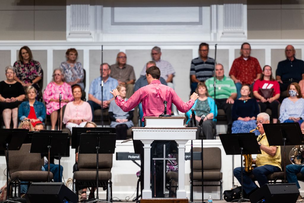 A male conductor leads a choir rehearsal with a group of adults in a church setting.