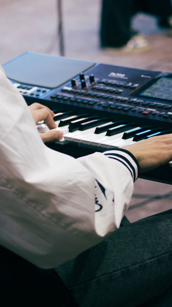 A musician wearing a white shirt plays an electronic keyboard indoors during the day.