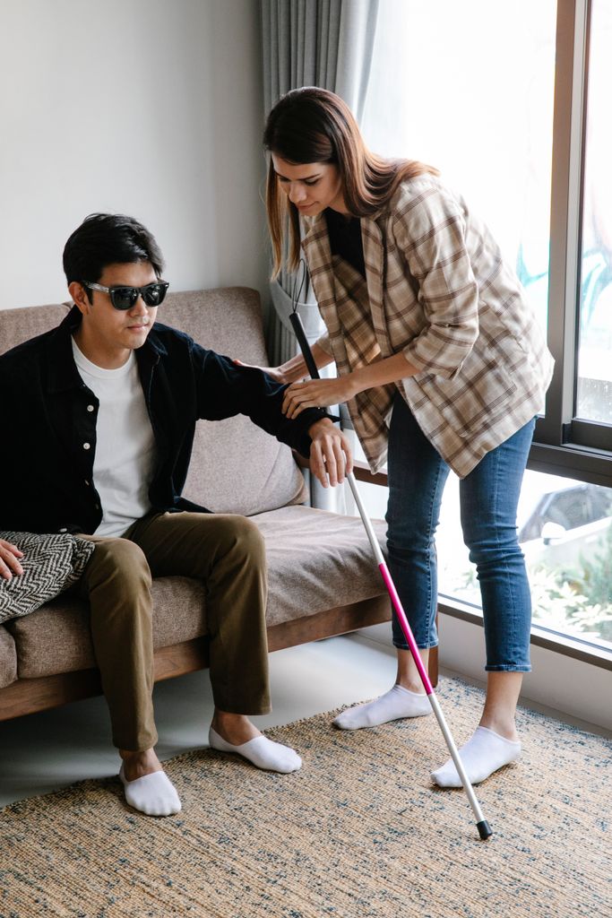 A woman provides guidance to a visually impaired man using a walking stick indoors on a cozy sofa.