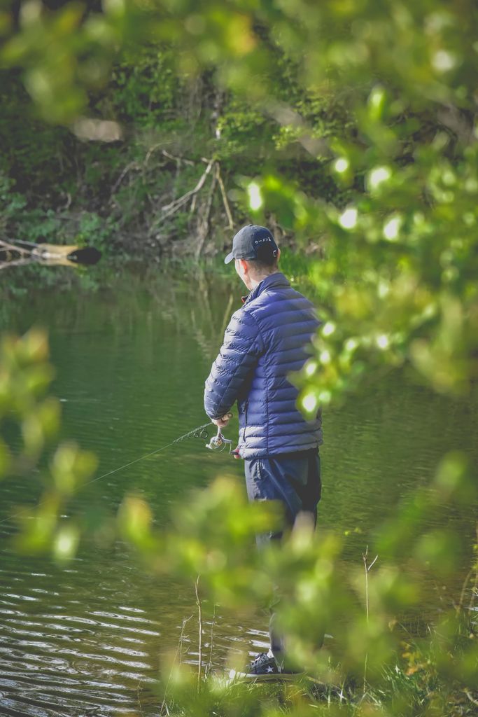 Back view of unrecognizable fisher in waterproof outerwear standing near river with fishing rod