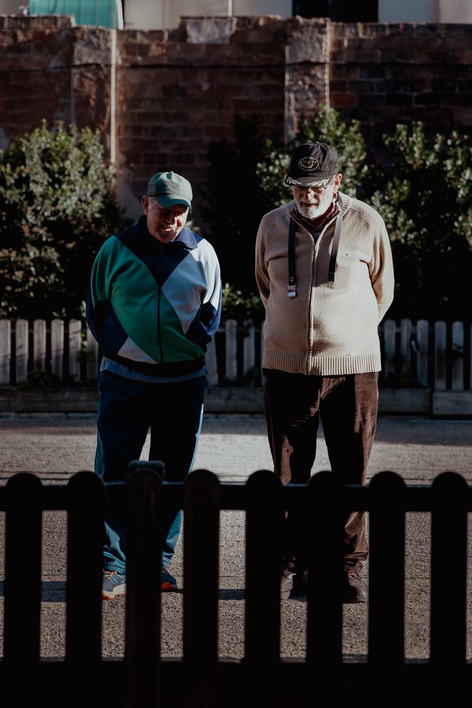 Two senior men walk together in a sunlit urban park in Barcelona, Spain.